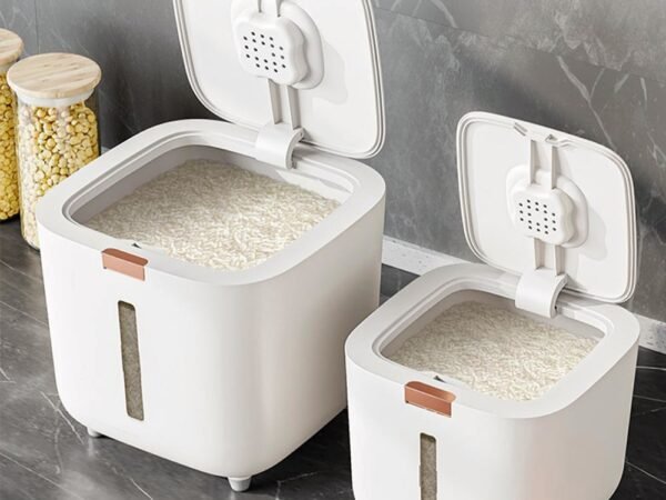 A white, modern airtight rice dispenser sitting on a kitchen counter, with the included measuring cup next to it.